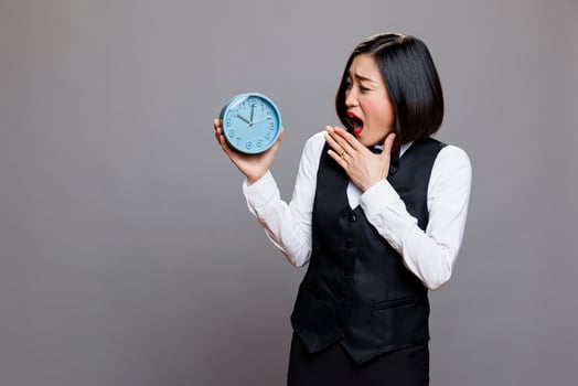 Sleepy asian waitress yawning and covering mouth with hand while looking at alarm clock. Tired young woman receptionist wearing restaurant uniform checking time while working late photo