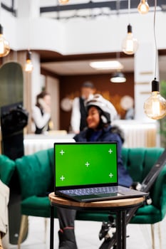 Detailed shot of digital device with isolated copyspace template placed on bench in lounge area of luxury ski mountain resort. Woman in wintersports gear at background of laptop with green screen. photo