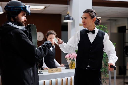 Friendly asian bellboy receiving cash tip from caucasian man with snow clothing at front desk of ski mountain resort. Male traveler thanking employee for assistance with luggage in hotel lobby. photo