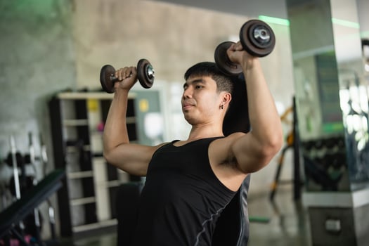 Asian man in a black tank top performs a seated dumbbell shoulder press in a modern gym. His muscular build and concentrated expression highlight his dedication to strength training. photo