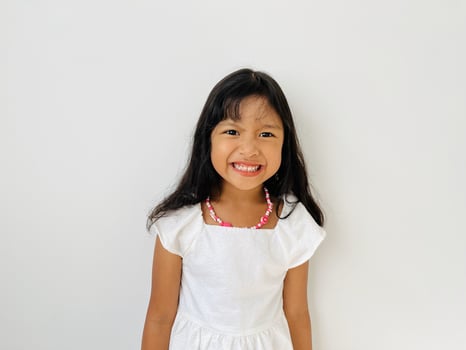 Portrait of a 5 year old Asian girl with shoulder length black hair wearing a white dress and necklace smiling broadly at the camera photo
