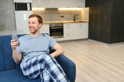A happy smiling caucasian guy, in casual stylish clothes, sits on the couch at home, holds a phone and a banking card in his hands, makes online payment for purchases, enters a card number photo