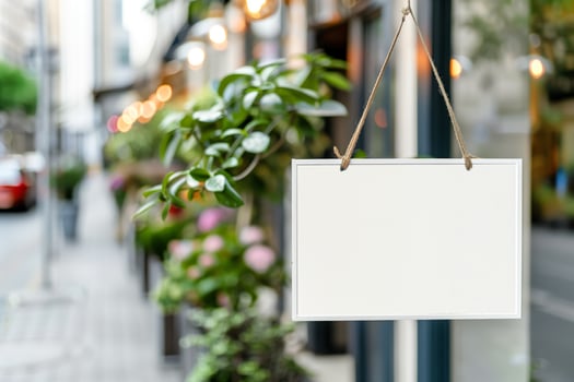 Blank white signboard hanging outside a shop on a city street, surrounded by green plants and urban decor. photo