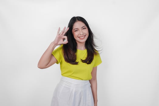 A happy young Asian woman in a yellow shirt and white skirt is giving okay good hand gesture, isolated by white background. photo
