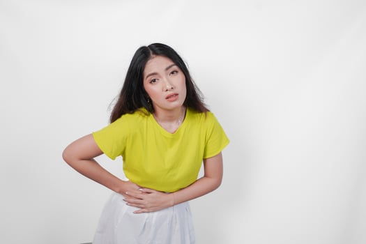 A sick Asian woman in a yellow shirt and white skirt is holding her stomach. She looks uncomfortable and is likely experiencing abdominal pain. photo