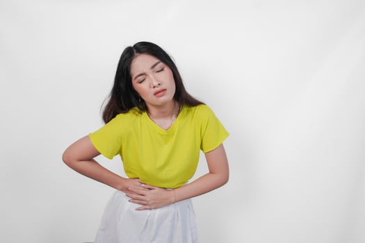 A sick Asian woman in a yellow shirt and white skirt is holding her stomach. She looks uncomfortable and is likely experiencing abdominal pain. photo