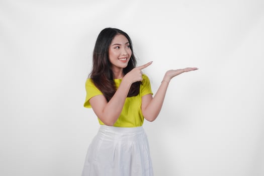 Excited young Asian woman wearing yellow shirt and white skirt pointing to the copy space beside her, isolated by white background. photo