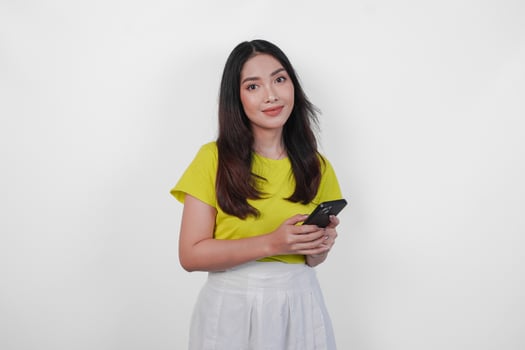 Cheerful Asian woman in a yellow shirt is holding a cell phone, smiling to the camera feeling happy, isolated by white background. photo