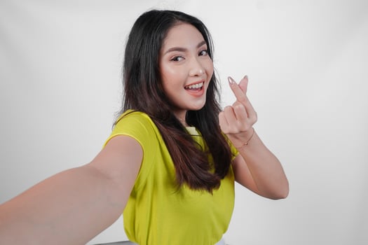 Lovely young Asian woman in a yellow shirt is smiling and showing finger heart gesture to the camera, isolated by white background. photo