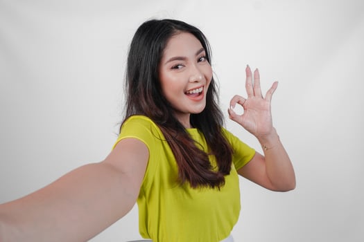 Attractive young Asian woman in a yellow shirt is taking a selfie and giving okay sign. Concept of happiness and positivity, as the woman is smiling and expressing approval. photo