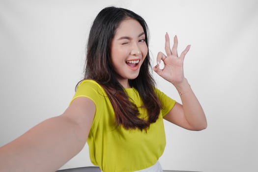 Attractive young Asian woman in a yellow shirt is taking a selfie and giving okay sign. Concept of happiness and positivity, as the woman is smiling and expressing approval. photo