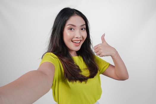 Cheerful young Asian woman in a yellow shirt is taking a selfie and giving thumb up approval sign isolated over white background. photo
