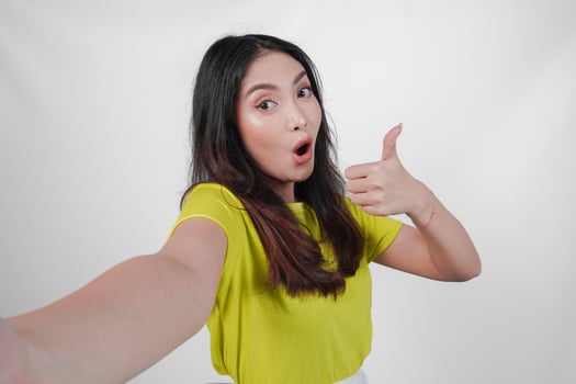 Cheerful young Asian woman in a yellow shirt is taking a selfie and giving thumb up approval sign isolated over white background. photo