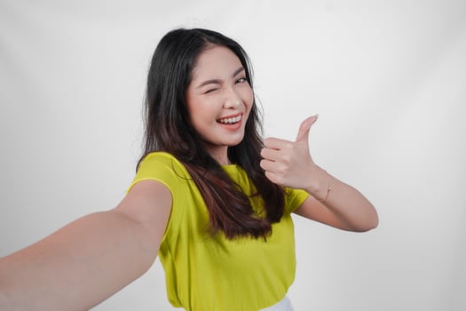Cheerful young Asian woman in a yellow shirt is taking a selfie and giving thumb up approval sign isolated over white background. photo