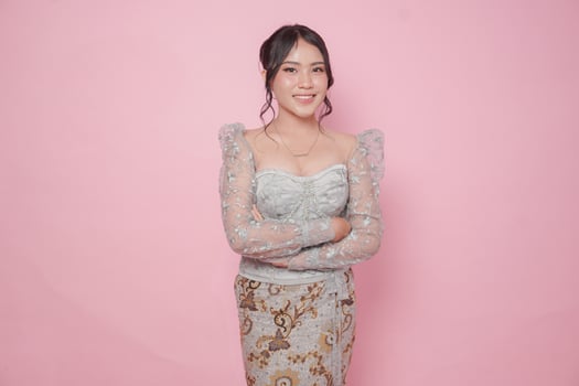 Confident smiling Asian woman wearing kebaya dress standing with arms folded, isolated over pink background. photo