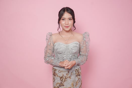 Beautiful young Indonesian woman wearing modern Kebaya dress standing and smiling at camera over isolated pink background. photo