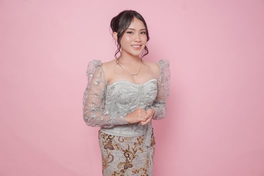 Beautiful young Indonesian woman wearing modern Kebaya dress standing and smiling at camera over isolated pink background. photo