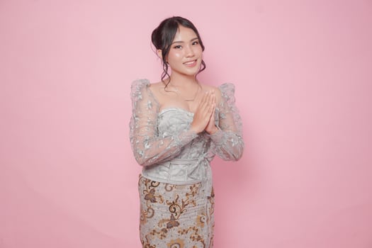 Elegant young Indonesian woman wearing modern kebaya dress gesturing greeting or namaste isolated over pink background. photo