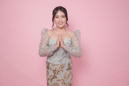 Elegant young Indonesian woman wearing modern kebaya dress gesturing greeting or namaste isolated over pink background. photo