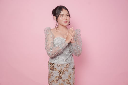 Elegant young Indonesian woman wearing modern kebaya dress gesturing greeting or namaste isolated over pink background. photo