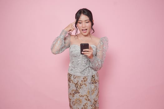 Cheerful Indonesian young woman wearing a modern kebaya dress holding smartphone and finding idea browsing internet over isolated pink background. photo