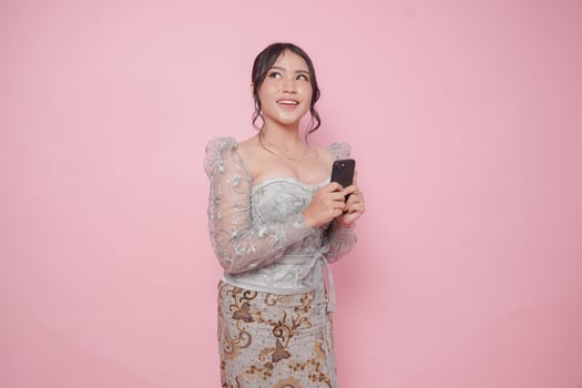 Indonesian young woman wearing a modern kebaya dress holding smartphone and looking at copy space upwards over isolated pink background. photo