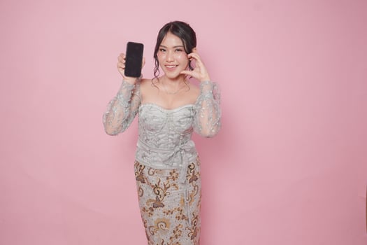 Joyful Indonesian young woman in a modern kebaya dress presenting copy space on smartphone and making heart shape gesture, isolated pink background. photo