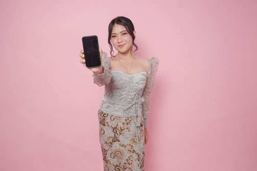 Smiling Indonesian young woman in a modern kebaya dress presenting copy space on smartphone, isolated pink background. photo