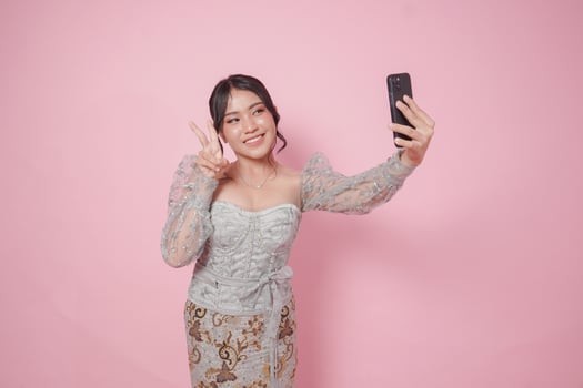Beautiful Indonesian woman wearing modern kebaya doing call pose while holding two fingers or peace sign isolated over pink background. photo