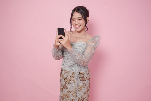 Cheerful Asian woman wearing modern kebaya using phone to text or browse internet, isolated by pink background. photo