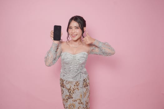 Excited Indonesian woman in modern kebaya dress gives thumbs up hand gesture of approval while showing copy space on her phone, isolated by pink background. photo