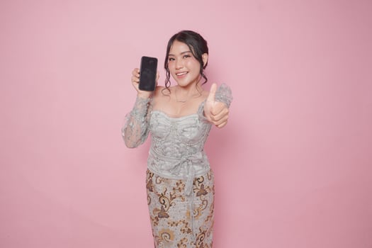 Excited Indonesian woman in modern kebaya dress gives thumbs up hand gesture of approval while showing copy space on her phone, isolated by pink background. photo