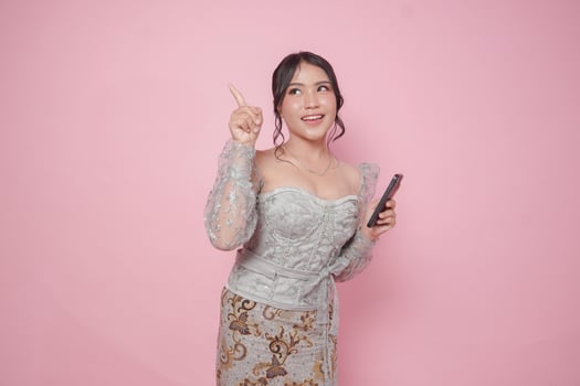 Joyful Asian woman wearing modern kebaya dress pointing at the copy space upside her while holding a smartphone, isolated by pink background. photo