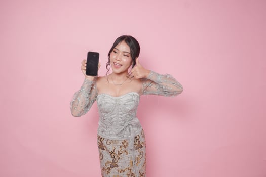 Excited Indonesian woman in modern kebaya dress gives thumbs up hand gesture of approval while showing copy space on her phone, isolated by pink background. photo