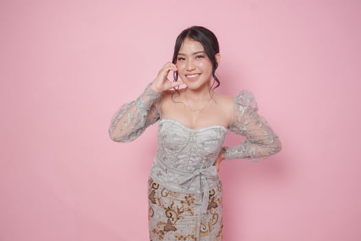 Lovely young Asian woman wearing modern kebaya dress making heart shape gesture on cheek, isolated by pink background. photo