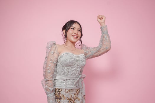 Beautiful Asian woman wearing modern kebaya dress gesturing oration with clenched fist hands isolated on pink background. photo