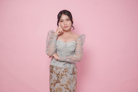 Thoughtful Asian woman wearing modern kebaya dress tilting her head, isolated by pink background. photo