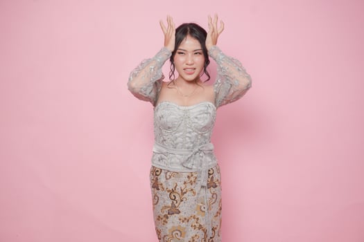 Young Asian woman wearing modern kebaya dress looking confused while holding her head, isolated by pink background. photo