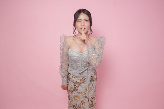 Young Asian woman wearing modern kebaya dress putting a finger in front of lips gesturing stay silent or stay quiet, isolated by pink background. photo
