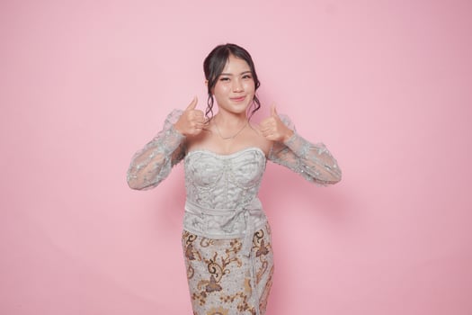 Excited Indonesian woman in modern kebaya dress gives thumbs up hand gesture of approval, isolated by pink background. photo