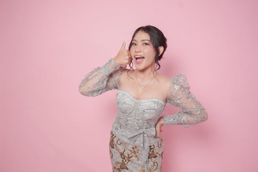 Excited Indonesian woman in modern kebaya dress gives thumbs up hand gesture of approval, isolated by pink background. photo