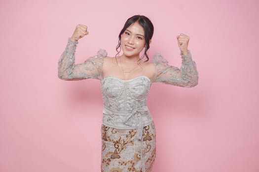 Confident Asian woman wearing modern kebaya dress showing strong gesture by lifting her arms and muscles over isolated pink background. photo