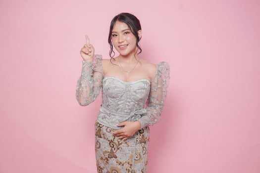 Beatiful Asian woman wearing modern kebaya dress pointing at the copy space upside her, isolated by pink background. photo