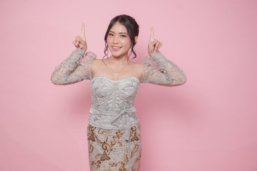 Beatiful Asian woman wearing modern kebaya dress pointing at the copy space upside her, isolated by pink background. photo
