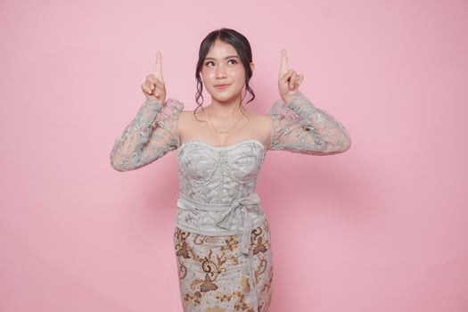 Beatiful Asian woman wearing modern kebaya dress pointing at the copy space upside her, isolated by pink background. photo