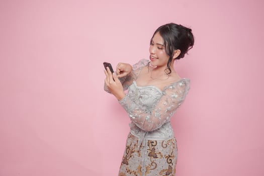 Cute Asian woman wearing modern kebaya dress pointing her smartphone, isolated by pink background. photo