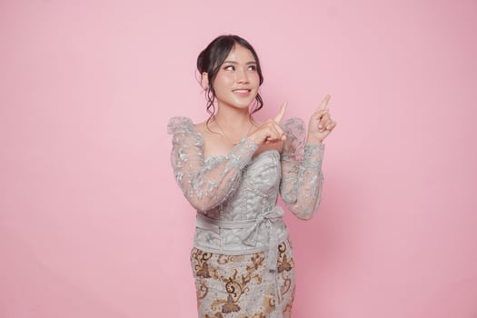 Joyful Asian woman wearing modern kebaya dress pointing at the copy space beside her, isolated by pink background. photo