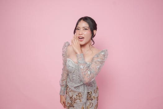 Young Asian woman wearing modern kebaya dress with hand on mouth telling secret rumor, whispering malicious talk conversation, isolated by pink background. photo