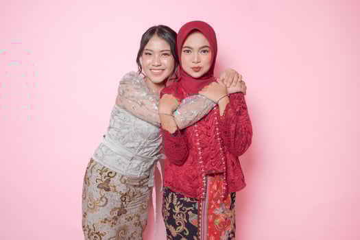 Happy two Asian young woman as close friends wearing modern kebaya and hugging each other showing friendly gesture, isolated over pink background. photo
