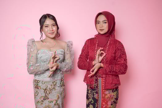 Two beautiful Asian women wearing modern kebaya dress doing meditation hand gesture greeting over isolated pink background. photo